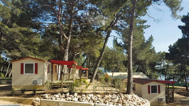 A row of small, single-story cabins with red roofs, surrounded by trees and a rocky garden area. - Victoria Mobilehome Camping Poljana (Mali Losinj)