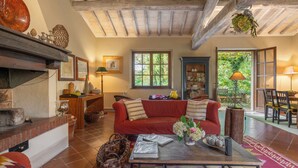 A cozy living room with a red sofa, a coffee table, and a fireplace. There are windows, a dining area, and a bookshelf. - Casa dell'Ambasciatore (Castiglione d'Orcia)