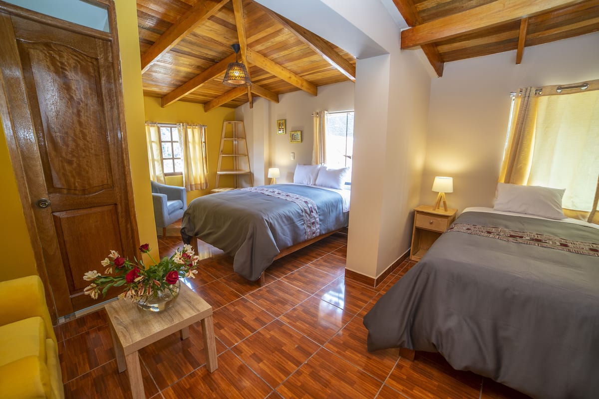 A bedroom with two beds, wooden flooring, a wooden ceiling, a small table with a vase of flowers, and a window with curtains.
