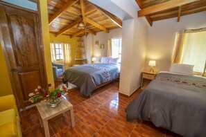 A bedroom with two beds, wooden flooring, a wooden ceiling, a small table with a vase of flowers, and a window with curtains. - San Isidro's House (Ollantaytambo)