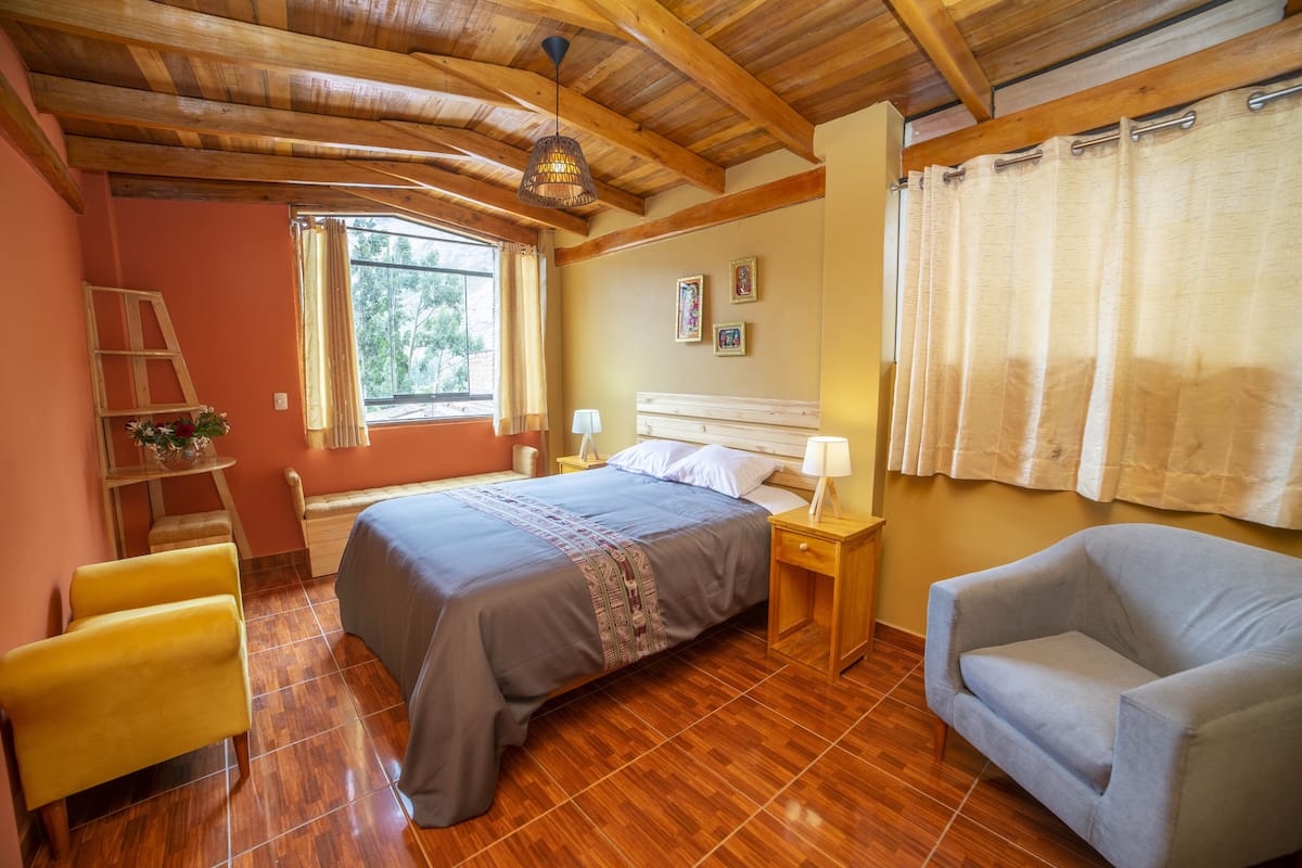 A bedroom with a wooden ceiling, a bed with a headboard, a nightstand, a chair, and a window with curtains.