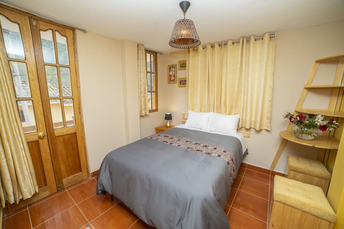 A bedroom with a bed, curtains, a nightstand, a lamp, and a small wooden shelf with flowers.