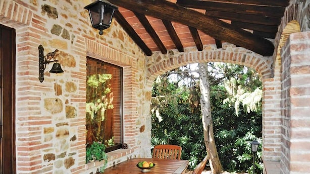 A stone-walled patio with a wooden table and chairs, a window with a view of greenery, and a lantern above the table. - Villa San Martino (Pomarance)
