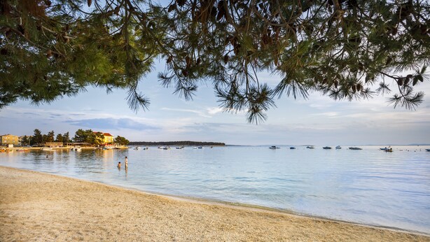 Una spiaggia con ciottoli, acqua calma, barche e edifici sullo sfondo.