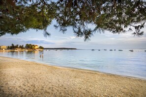 A beach with pebbles, calm water, boats, and buildings in the background. - Victoria Mobilehome Camping Padova (Rab)