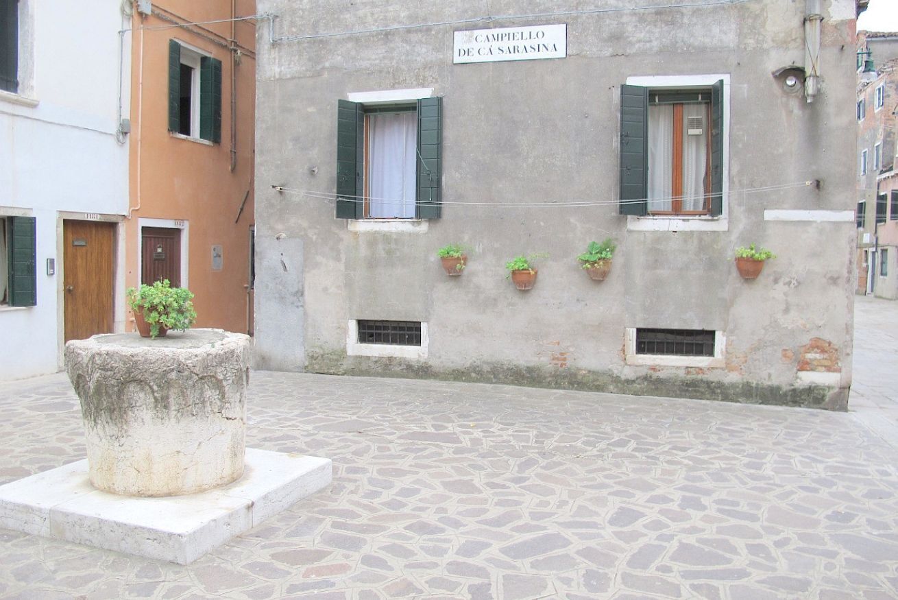 Ein Innenhof mit Steinboden, einem Steinsockel mit einer Pflanze und einem Gebäude mit der Aufschrift 'CAMPIELLO DE CÁ SARASINA'.