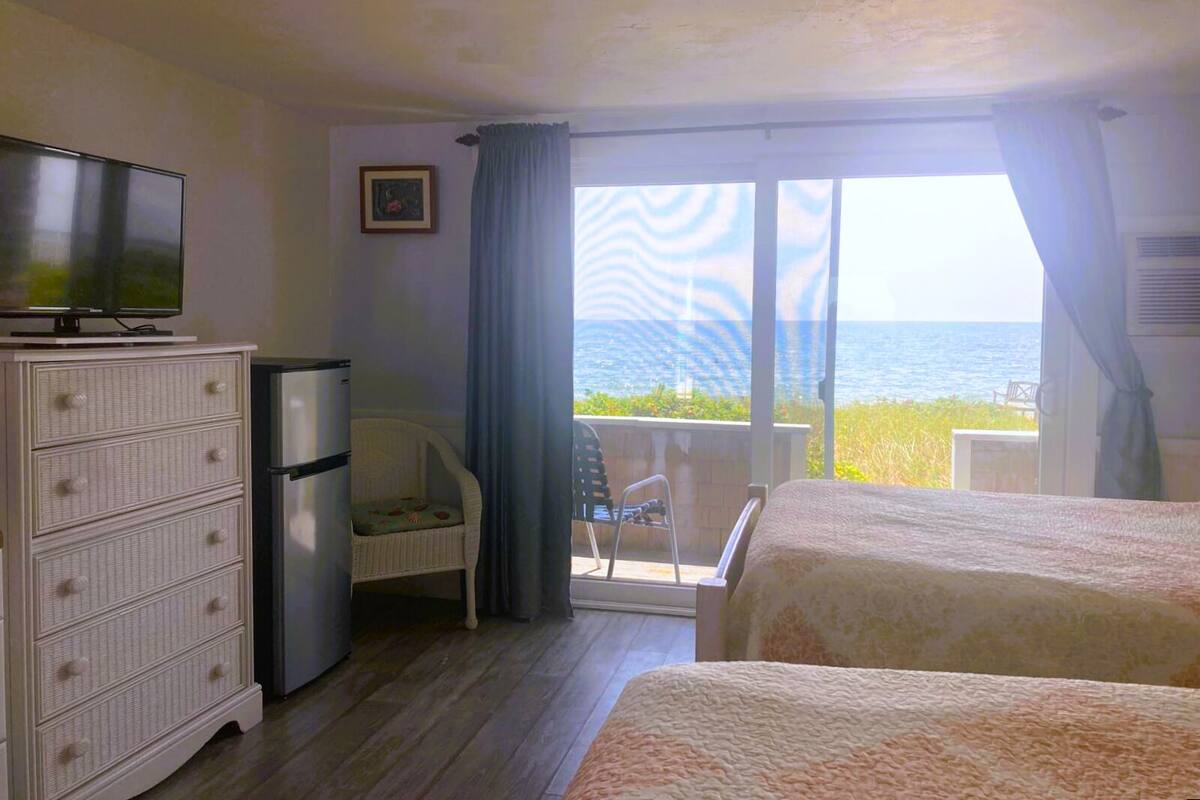 A hotel room with a bed, a TV, a dresser, and a balcony with a view of the sea.