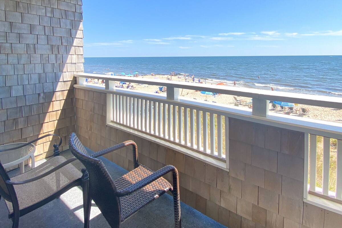 A covered outdoor area with a view of the beach, featuring two wicker chairs and a table.