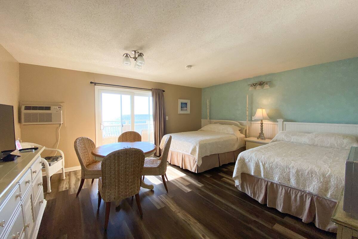 A hotel room with two beds, a dining table with chairs, a desk with a computer, and a window with a view of the ocean.