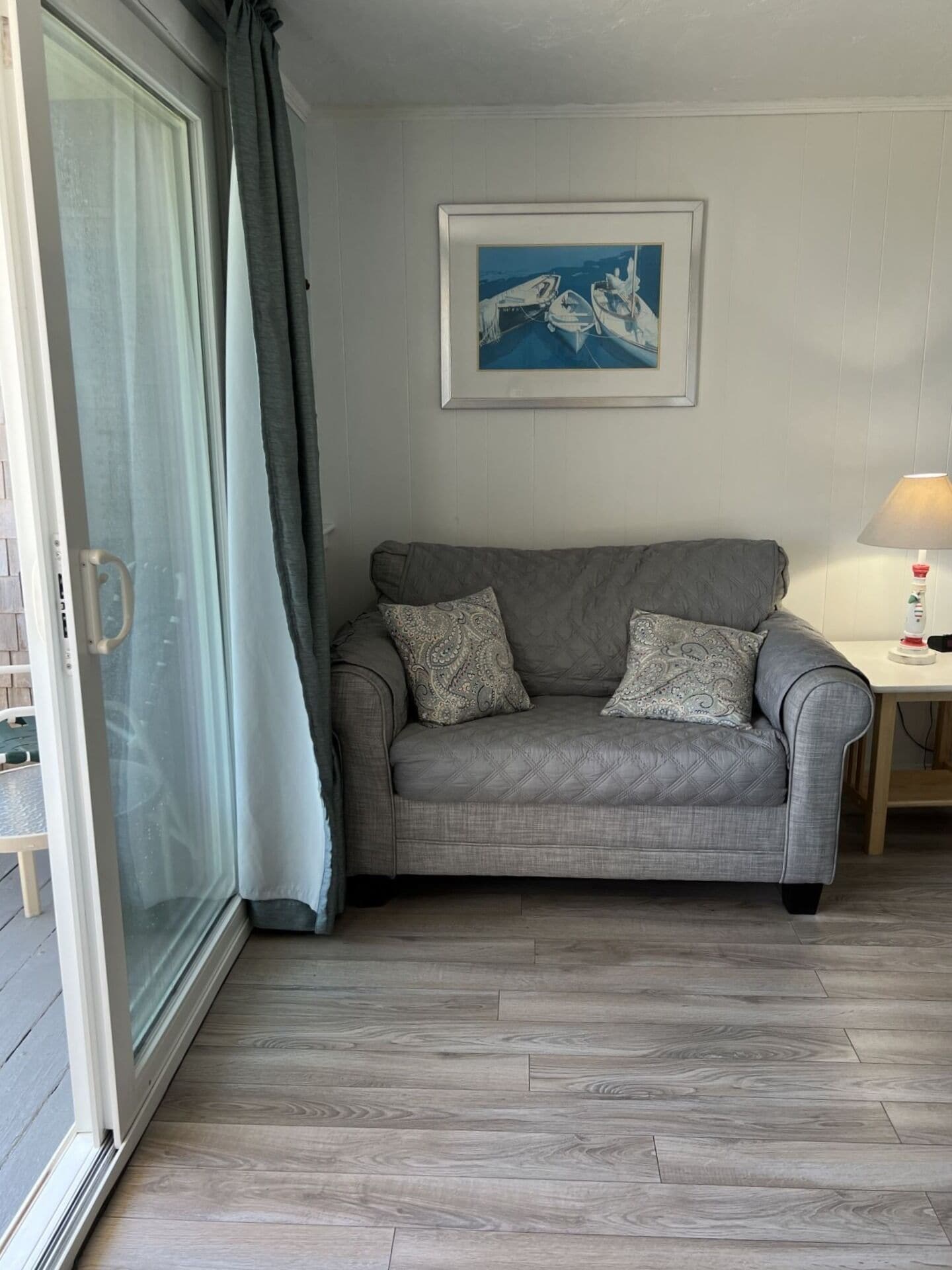 A grey sofa with patterned cushions, a framed artwork above it, a small table with a lamp, and a sliding glass door with curtains.