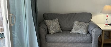 A grey sofa with patterned cushions, a framed artwork above it, a small table with a lamp, and a sliding glass door with curtains.