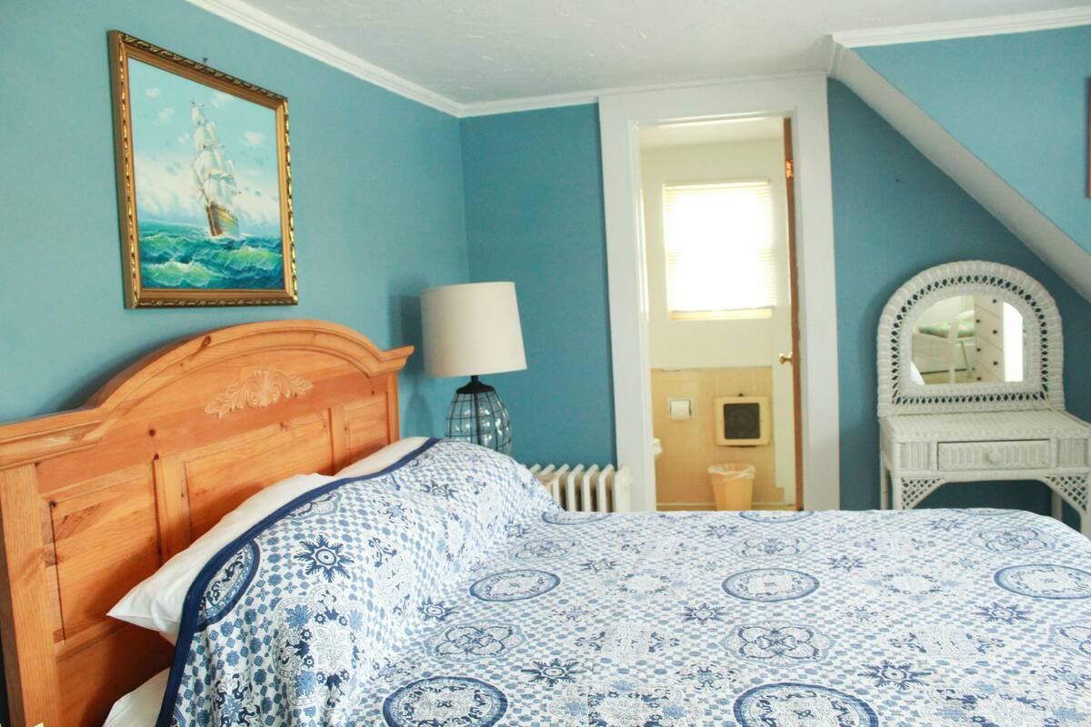 A bedroom with a wooden headboard, a patterned bedspread, a framed picture of a ship, a bedside lamp, and a dresser with a mirror.