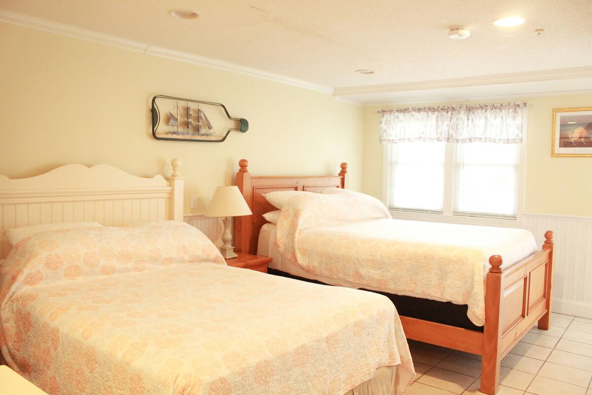 A bedroom with two single beds, a nightstand with a lamp, a framed picture on the wall, and a window with a floral curtain.