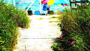 Un sentier en bois mène à une plage où se trouvent un parasol coloré et une chaise.