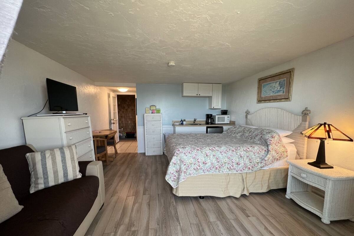 A bedroom with a bed, a bedside table with a lamp, a dresser, a television on a stand, a kitchenette with a microwave and coffee maker, and a door leading to a bathroom.
