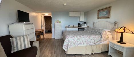 A bedroom with a bed, a bedside table with a lamp, a dresser, a television on a stand, a kitchenette with a microwave and coffee maker, and a door leading to a bathroom.