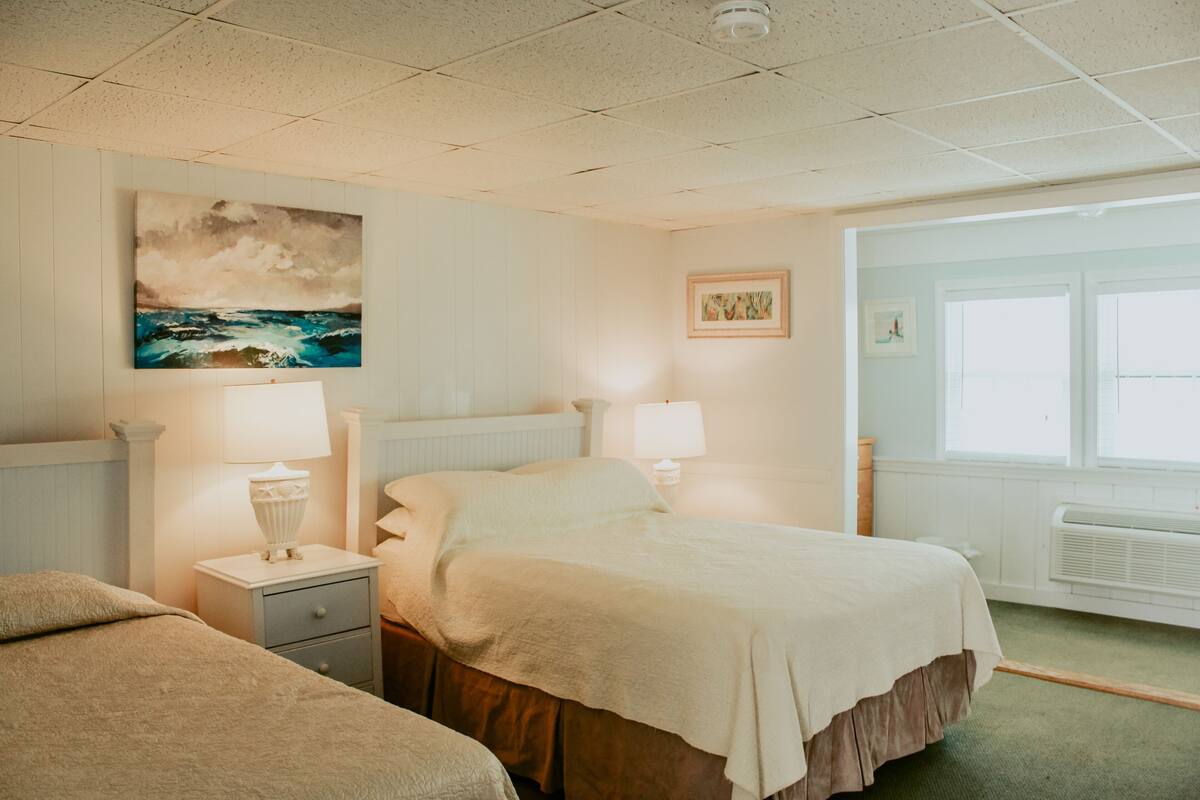 A hotel room with a bed, two bedside tables with lamps, a window with a view, and a painting on the wall.