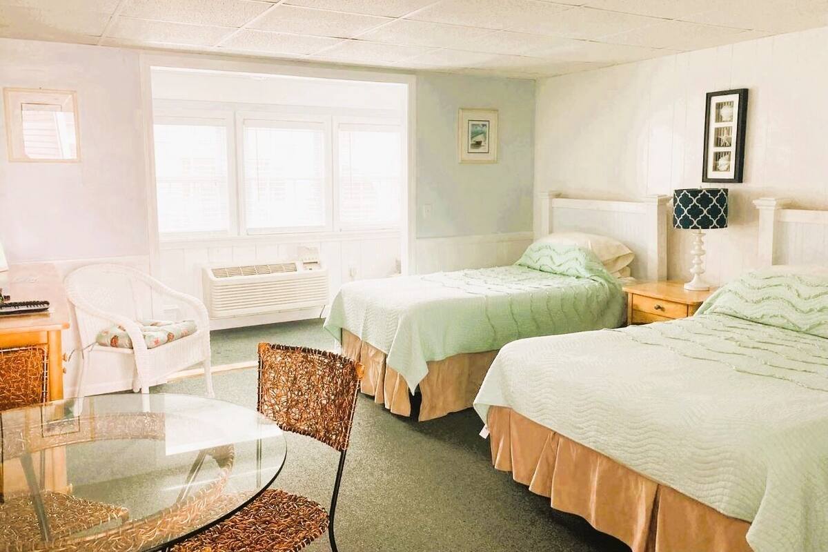 A hotel room with two beds, a chair, a table, and a window.