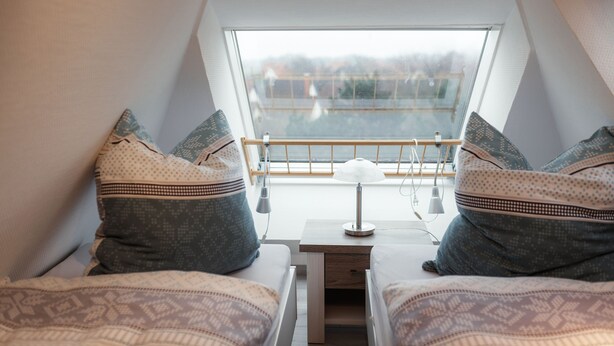 Ein gemütlicher, dreieckiger Raum mit zwei Betten, gemusterten Textilien, einem kleinen Tisch mit einer Lampe und Blick auf einen Balkon mit Geländer.
