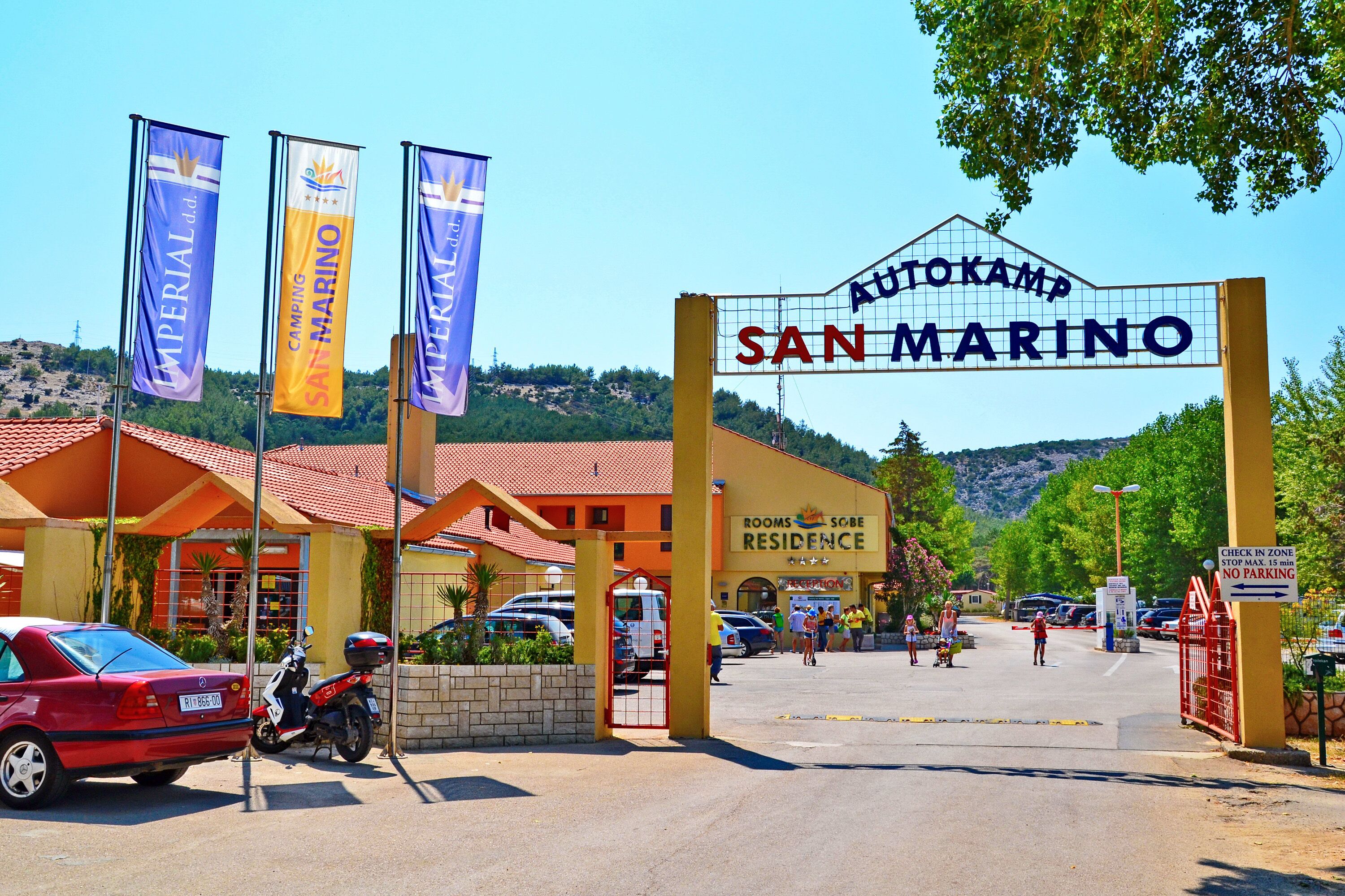 Entrada de un hotel con un cartel que dice "Autokamp San Marino" y banderas de "Imperial" y "Camping San Marino".