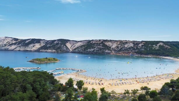 En kustnära strand med klart vatten, en sandstrand och ett bergigt landskap i bakgrunden.
