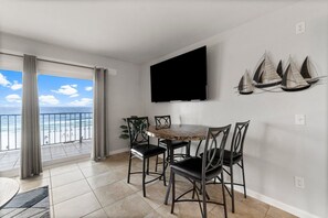 A dining area with a round table and four chairs, a flat-screen TV mounted on the wall, and a view of the beach through large windows.
