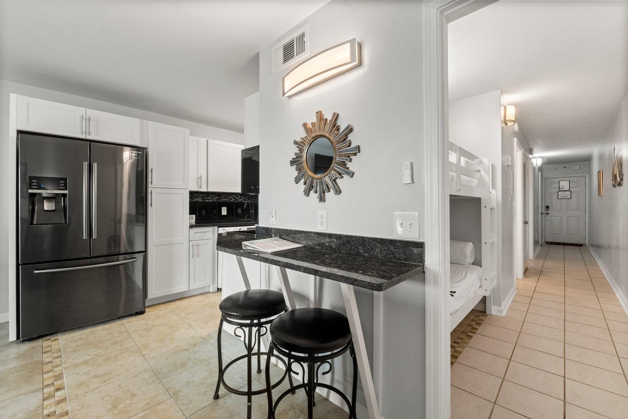 A modern kitchen with white cabinets, a stainless steel refrigerator, a granite countertop, and two black bar stools.