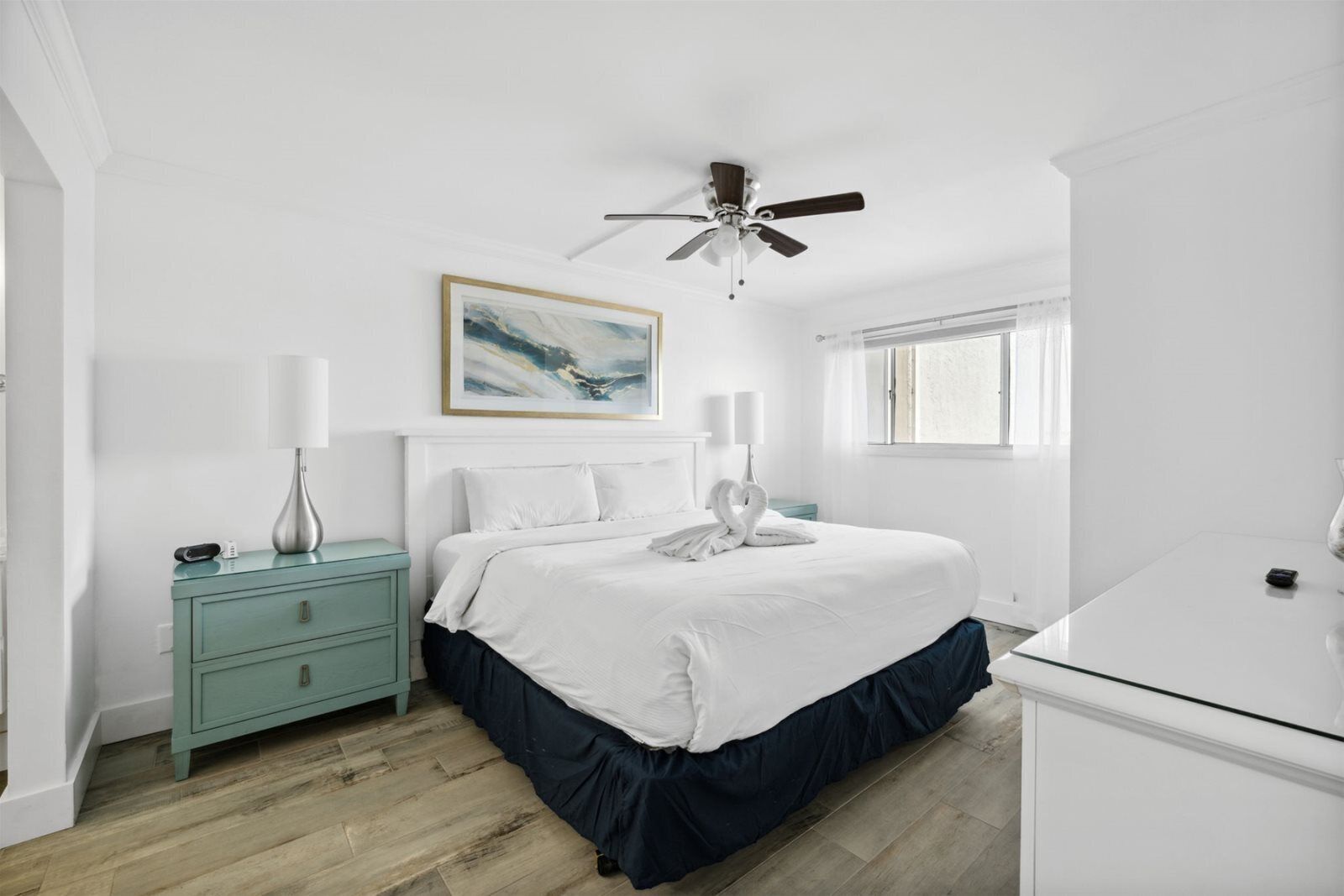 A bedroom with a bed, bedside tables, a ceiling fan, a window, and a painting on the wall.