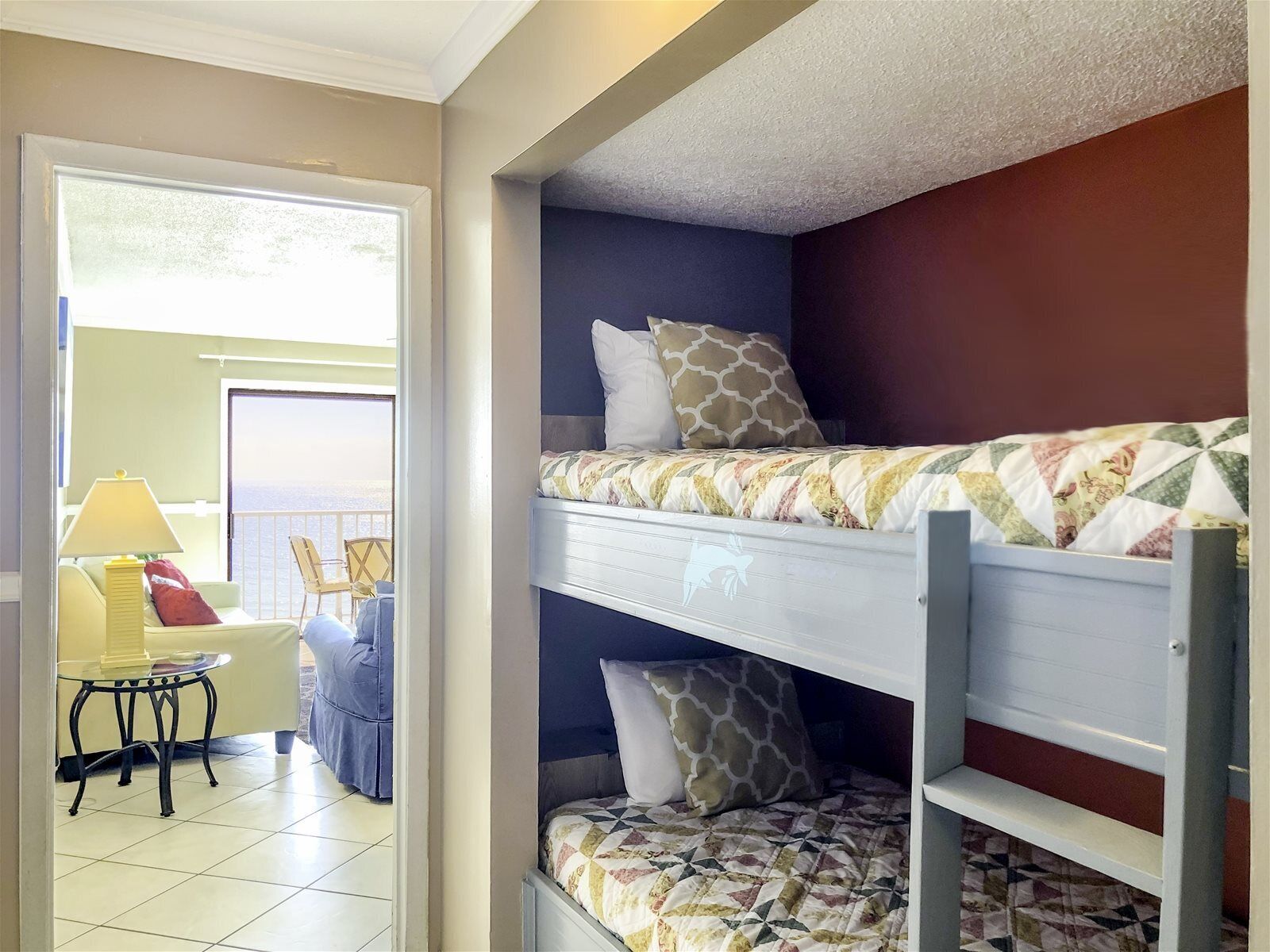 A room with a bunk bed, a small table with a lamp, and a view of the ocean through a door.