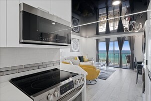 Cozinha moderna com forno embutido e fogão, área de jantar com uma cadeira amarela e varanda com vista para o oceano.