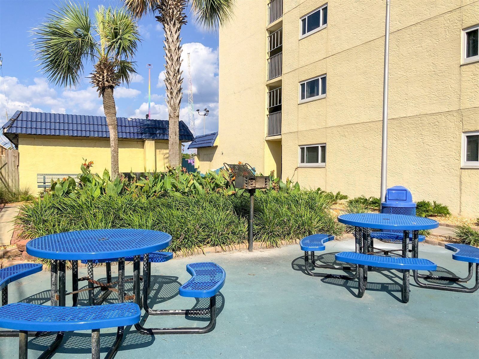 Un espace extérieur aménagé en terrasse, avec des tables et des bancs bleus, entouré de verdure et bordé d’un bâtiment doté de balcons.