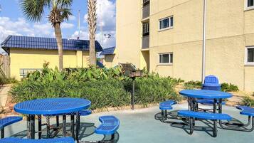 Un espace extérieur aménagé en terrasse, avec des tables et des bancs bleus, entouré de verdure et bordé d’un bâtiment doté de balcons.