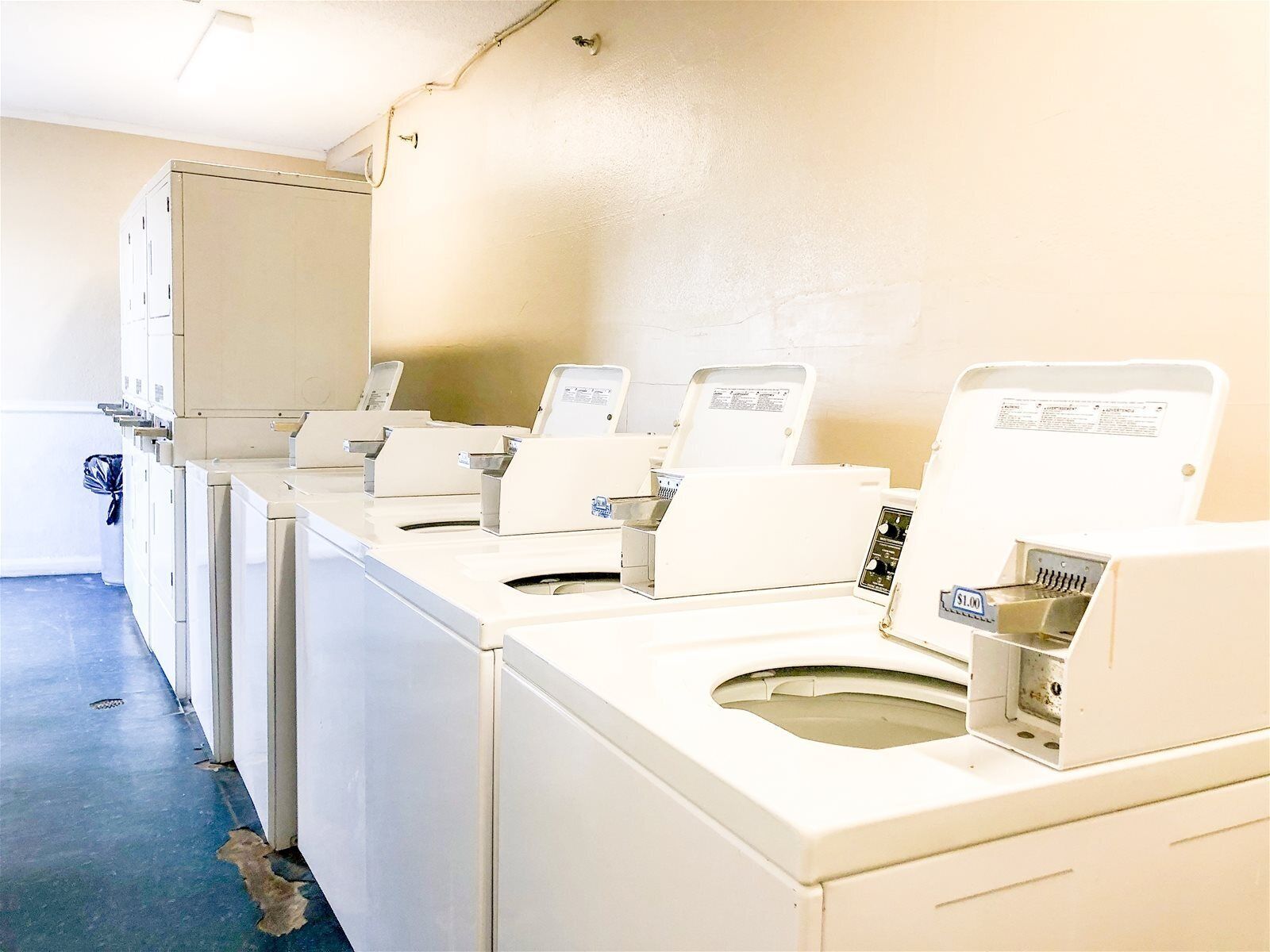 A row of white laundry machines in a room with a blue floor and beige walls.