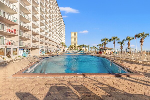Un espace piscine d’hôtel comprenant une grande piscine, des chaises longues, des palmiers et un grand immeuble en arrière-plan.