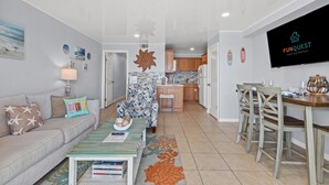 A modern living room with a sofa, coffee table, and TV. There is a kitchen area with cabinets and a dining table. - The Summit 1409 (Panama City Beach)