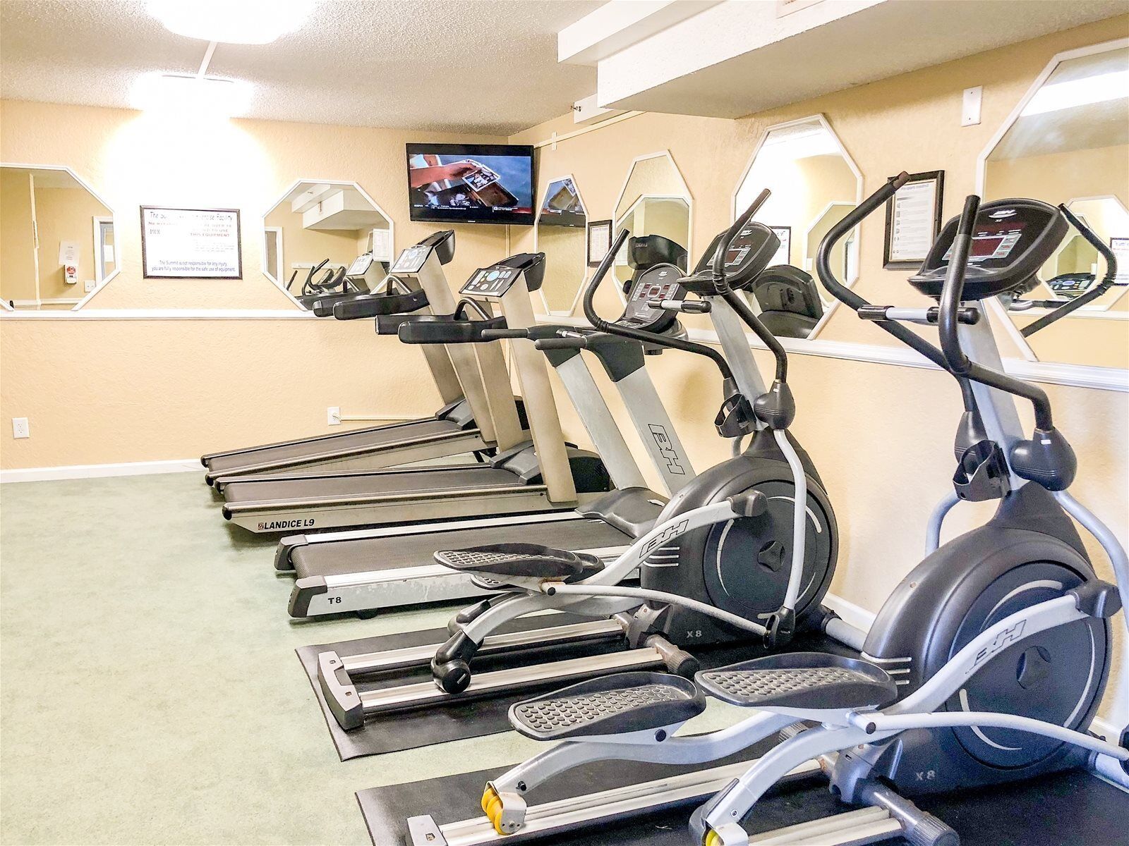 A gym with multiple treadmills and an elliptical machine, a TV on the wall, and framed certificates on the wall.