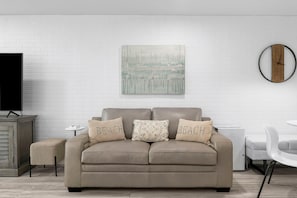A modern living room with a beige sofa, a small side table, a flat-screen TV, a decorative wall piece, and a round clock.