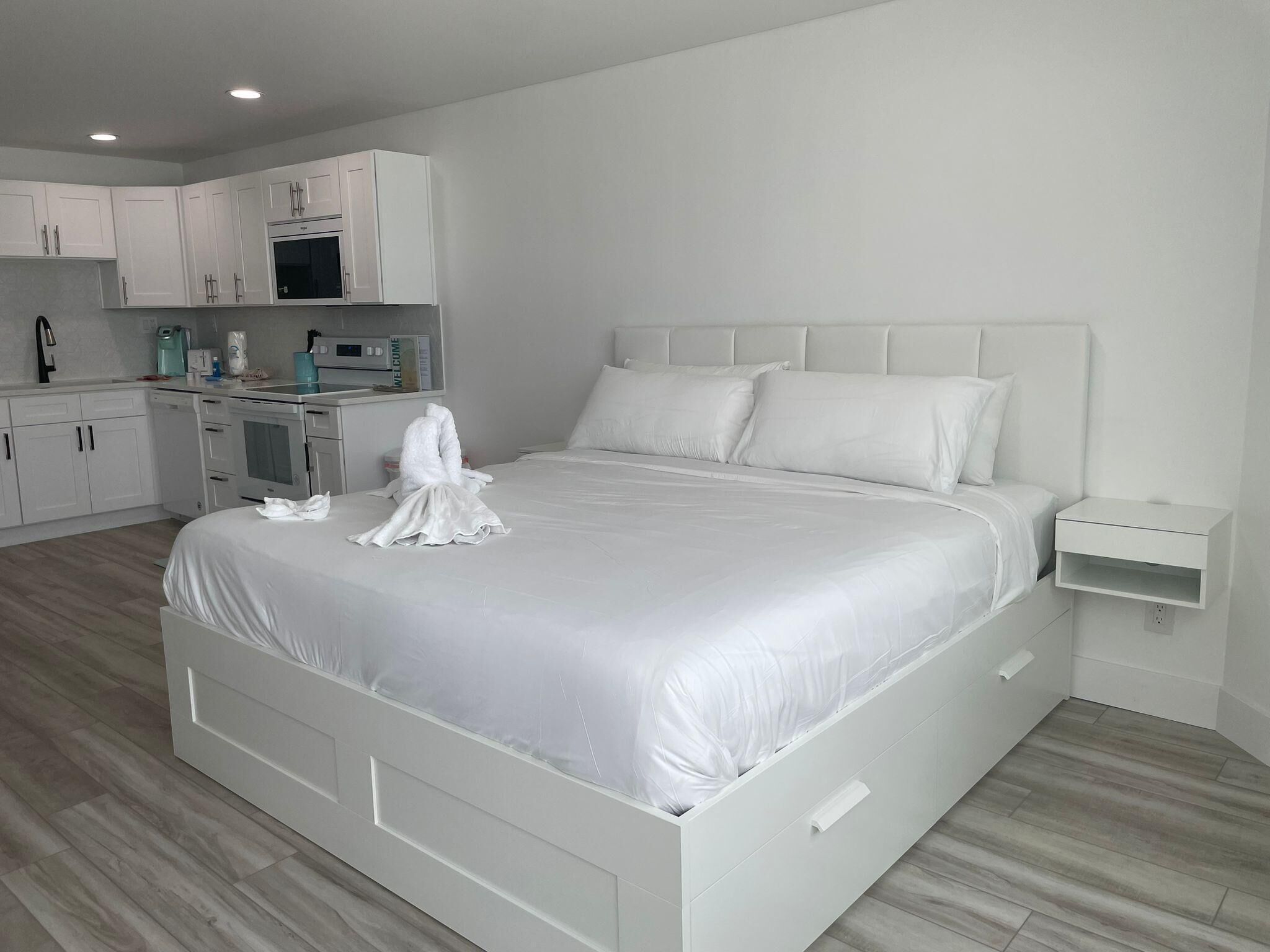 Quarto de hotel moderno com uma cama grande, uma pequena cozinha e uma decoração em tons de branco.