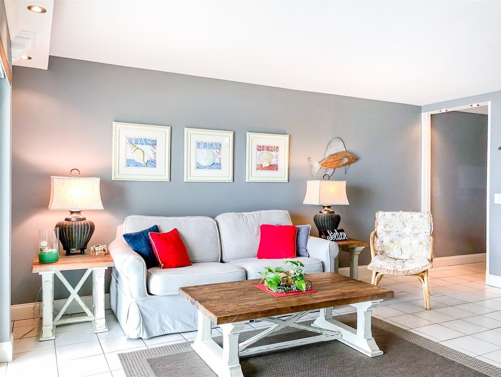 A living room with a white sofa, red and blue cushions, a wooden coffee table, and two lamps on side tables. There are three framed pictures on the wall.