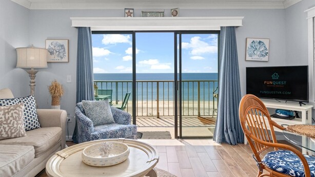 Een woonkamer met een bank, fauteuil en een televisie. Er is uitzicht op het strand en de oceaan door grote glazen deuren.
