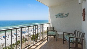 A balcony with a view of the beach, two chairs, a table, and a decorative wall piece.