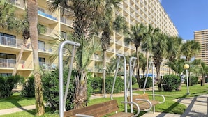 A row of wooden benches with metal armrests, a palm tree, and a building with balconies in the background. - The Summit 104 (Panama City Beach)