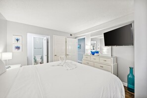 Ein Hotelzimmer mit einem großen Bett, einem an der Wand befestigten Fernseher, einem Schminktisch und einem durchgehenden Türspalte erkennbaren Badezimmer.