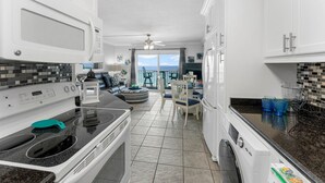 A modern kitchen with white cabinets, a microwave, stove, and a washing machine. There is a dining area with a table and chairs, and a view of the ocean through large windows. - The Summit 1130 (Panama City Beach)