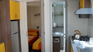 A compact kitchen with a refrigerator, sink, stove, and a bed with a colorful blanket.
