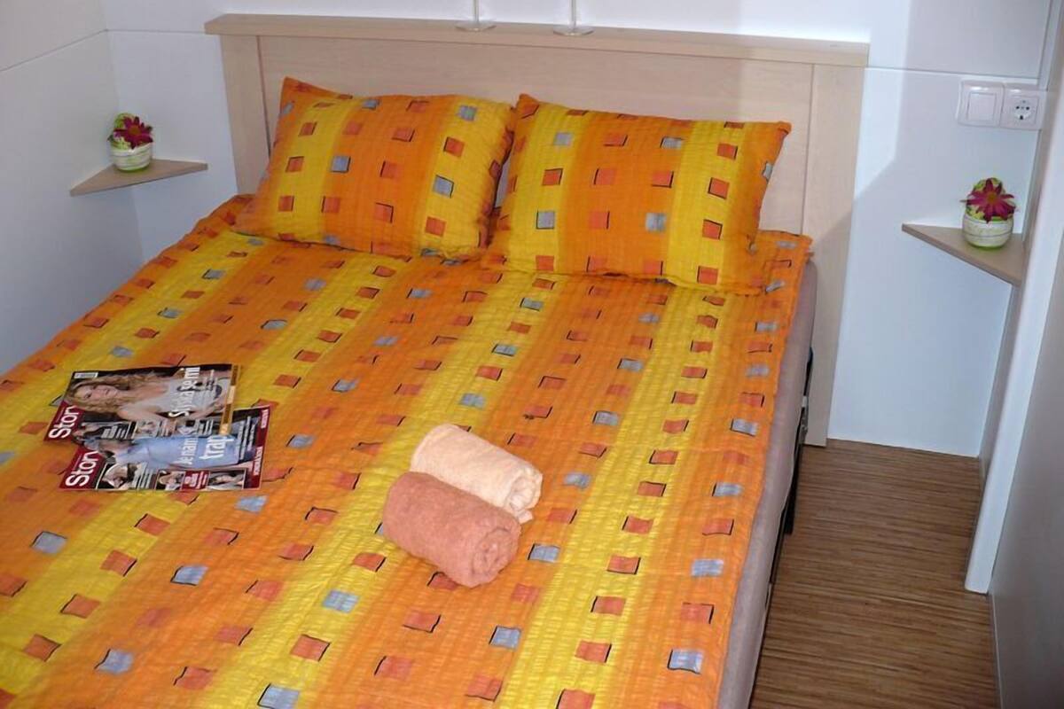 Un lit bien fait, recouvert d’une couvre-lit à motifs, avec deux oreillers, une serviette enroulée et un magazine posés dessus. Deux petites tables de chevet sont disposées de chaque côté du lit, ornées de quelques objets décoratifs.