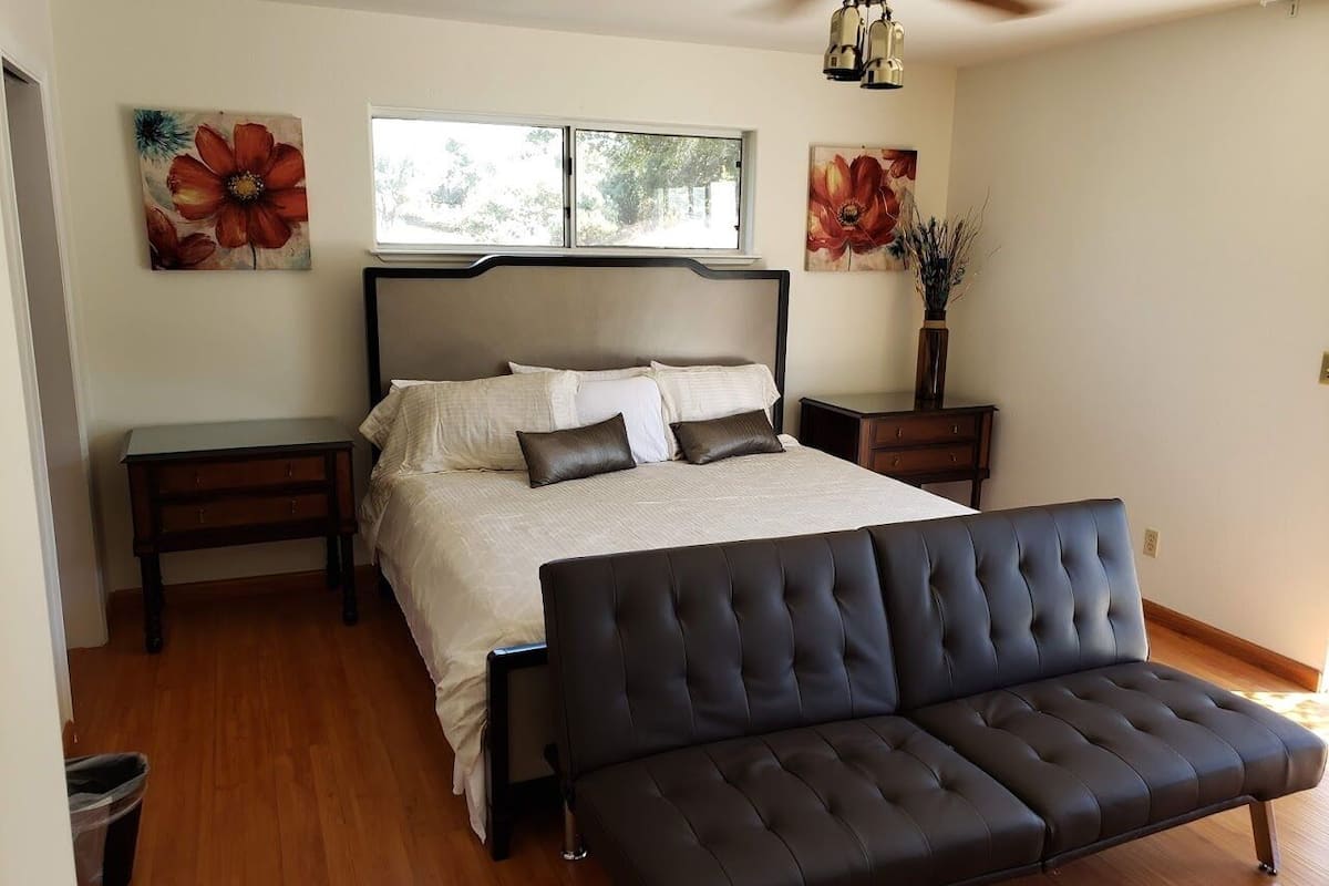 Un dormitorio con cama, mesitas de noche, un sofá, ventilador de techo y obras de arte en las paredes.
