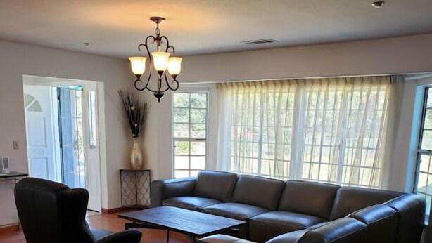 A spacious living room with a dark leather sofa, a black armchair, and a wooden coffee table. There is a chandelier hanging from the ceiling, and large windows with sheer curtains allow natural light to fill the room.