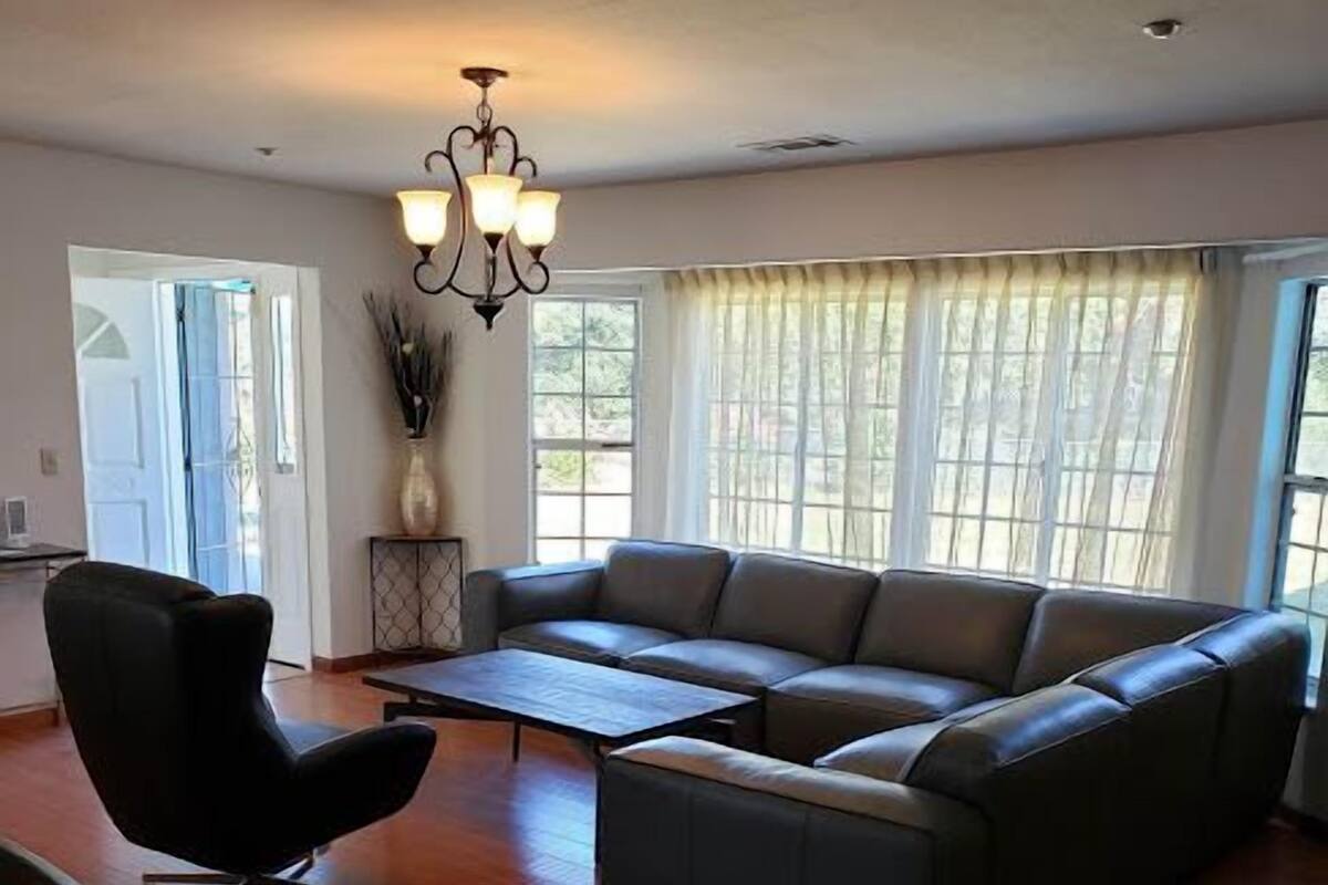 Amplia sala de estar con un sofá de cuero oscuro, un sillón negro y una mesa de centro de madera. Un candelabro cuelga del techo, y grandes ventanas con cortinas translúcidas dejan entrar abundante luz natural.
