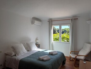 Une chambre à coucher avec un lit, des oreillers, une table de chevet avec une lampe, une fenêtre avec des rideaux et une chaise.